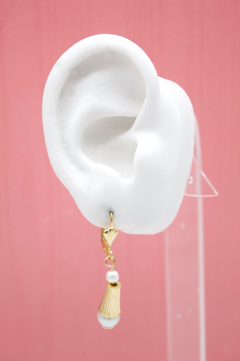 A white mannequin ear displays a gold dangle earring featuring a small pearl and a ribbed cone charm against a pink background.
