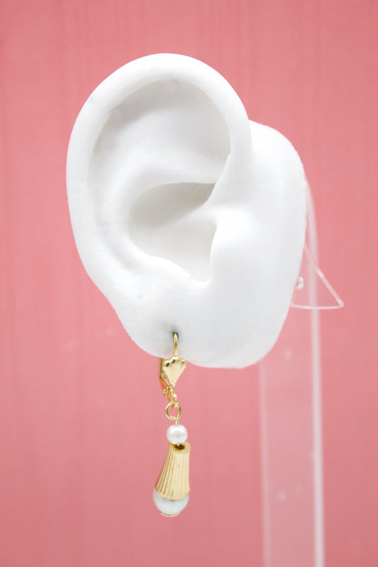 A white mannequin ear displays a gold dangle earring featuring a small pearl and a ribbed cone charm against a pink background.