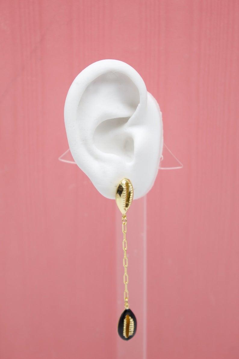 A white mannequin ear displays a gold-plated cowrie shell dangle earring against a pink background.