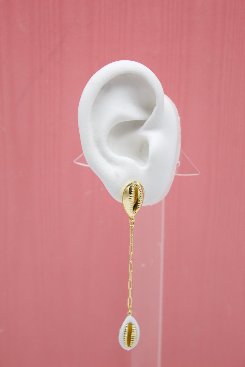 A white plaster ear model displays a gold cowrie shell dangle earring with a delicate chain against a pink background.