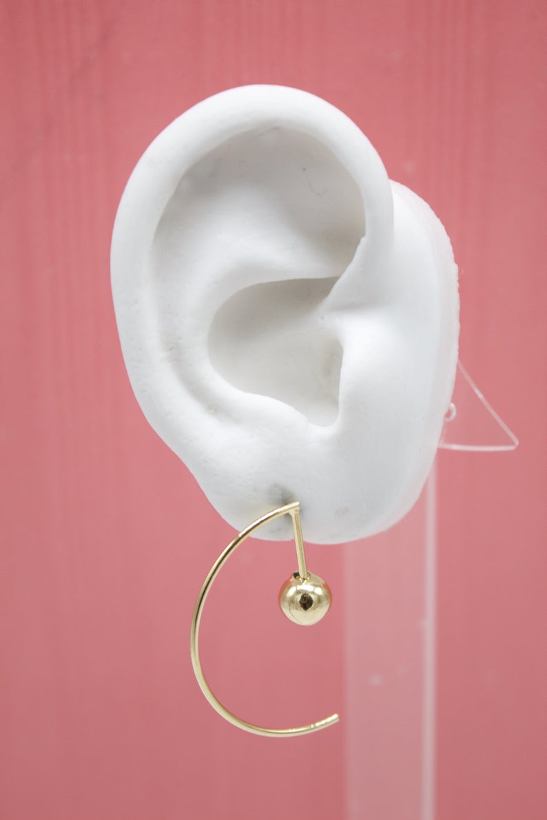 A minimalist gold hoop earring featuring a delicate dangling sphere hangs from a white mannequin ear against a solid pink backdrop.
