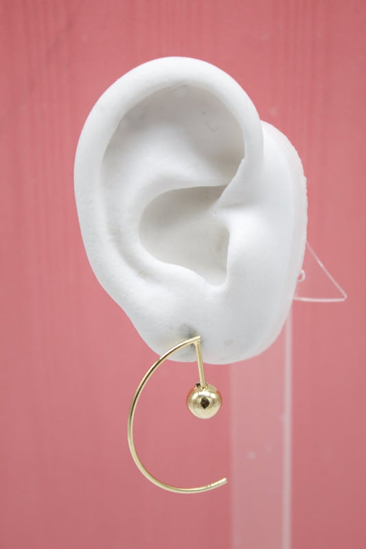 A minimalist gold hoop earring featuring a delicate dangling sphere hangs from a white mannequin ear against a solid pink backdrop.