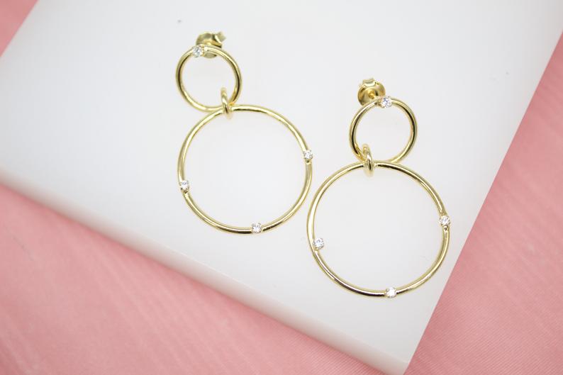A pair of elegant gold double hoop earrings embellished with sparkling cubic zirconia stones rests on a white display surface.