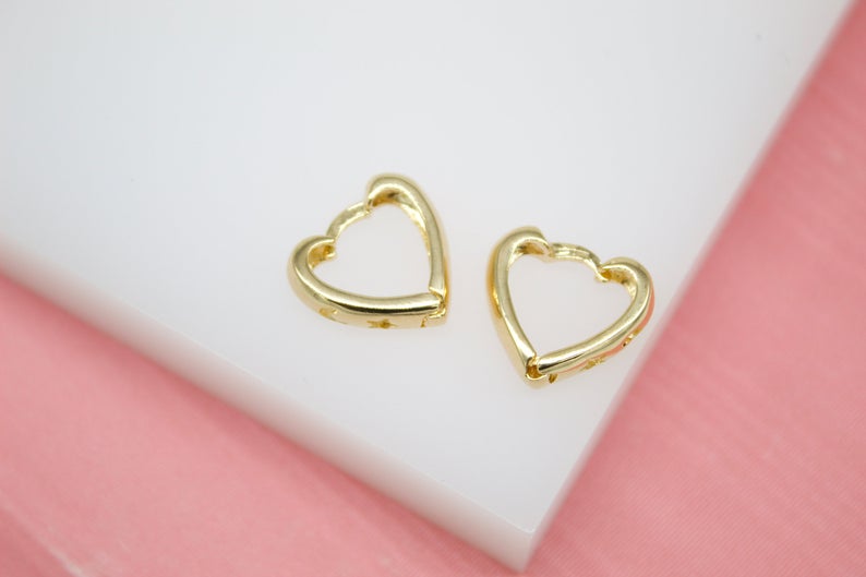 A pair of shiny gold heart-shaped hoop earrings rests on a white display surface against a soft pink background.