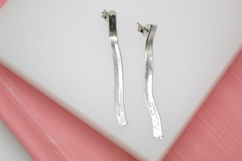 A pair of dangling silver herringbone chain earrings rests on a white display against a pink background.