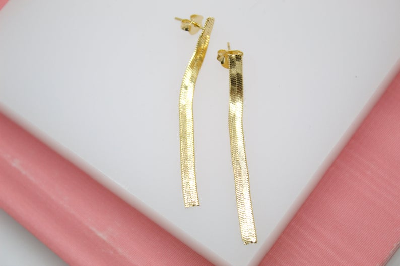 A pair of elegant gold herringbone chain drop earrings rests against a white and pink display surface.