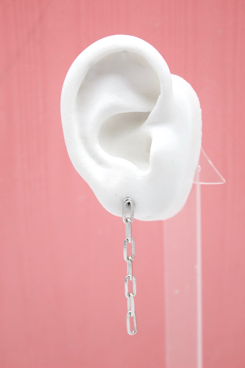 A white mannequin ear displays a dangling silver chain link earring against a pink background.