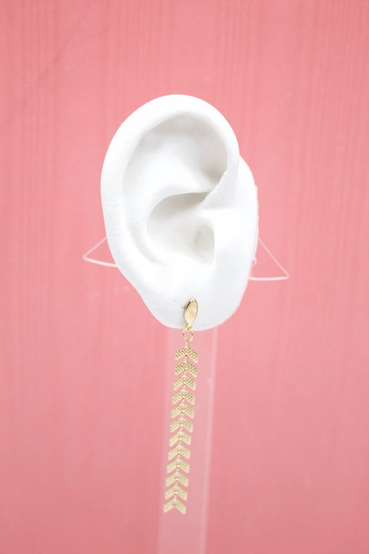 A white mannequin ear displays a long, gold chevron dangle earring against a pink background.