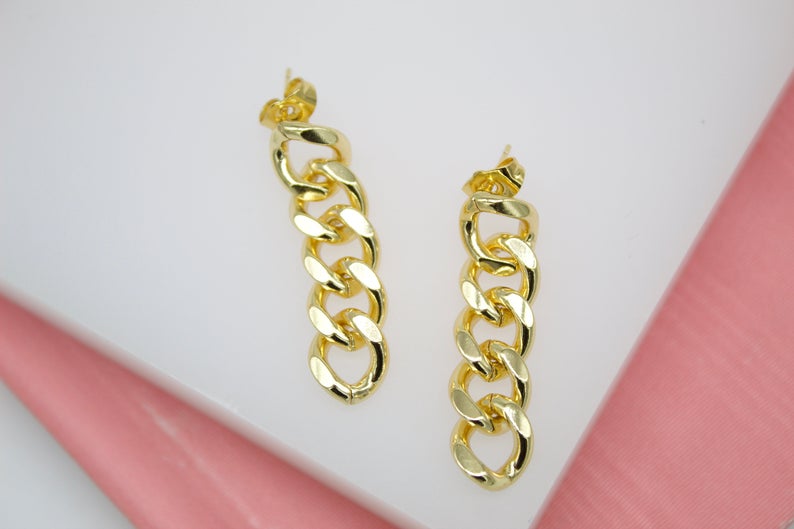 A pair of polished gold chain link drop earrings rests against a white and pink display surface.