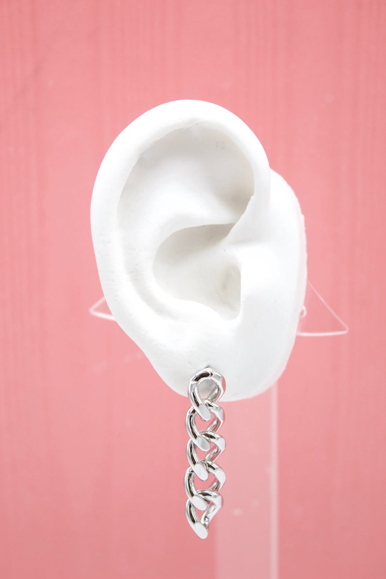 A white mannequin ear displays a dangling silver chain link earring against a pink background.