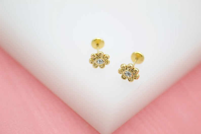 A pair of gold flower stud earrings with sparkling clear cubic zirconia centers rests on a white display against a soft pink background.