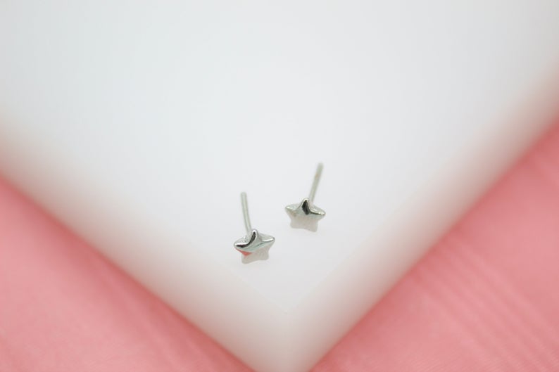 A pair of shiny silver star stud earrings rests on a white surface against a soft pink background.
