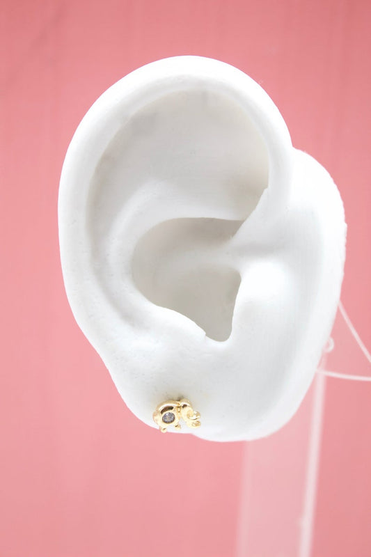 A white mannequin ear displays a cute gold elephant stud earring on a pink background.
