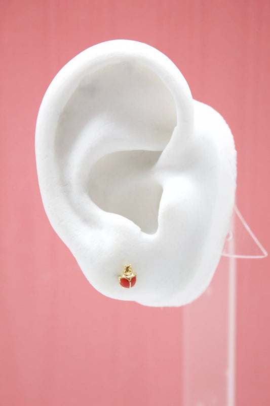 A white ear model displays a gold and red ladybug stud earring against a pink background.