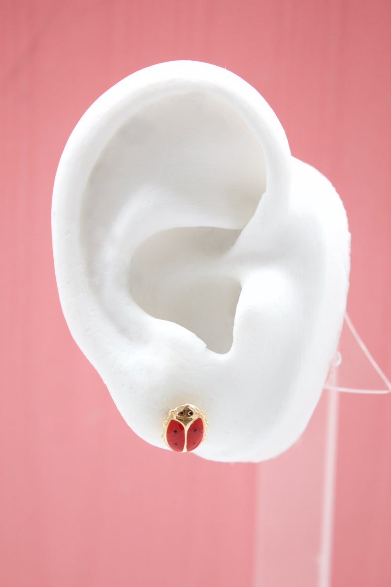A white mannequin ear displays a cute gold ladybug stud earring with red enamel wings against a solid pink backdrop.