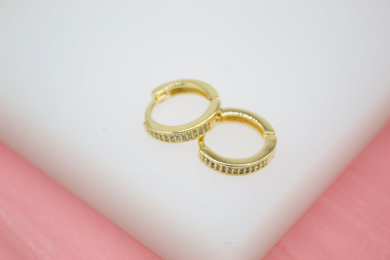 A pair of sparkling gold huggie hoop earrings rests delicately on a white and pink surface.