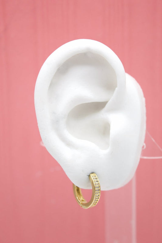 A white mannequin ear displays a sparkling gold huggie hoop earring encrusted with small clear crystals against a pink background.