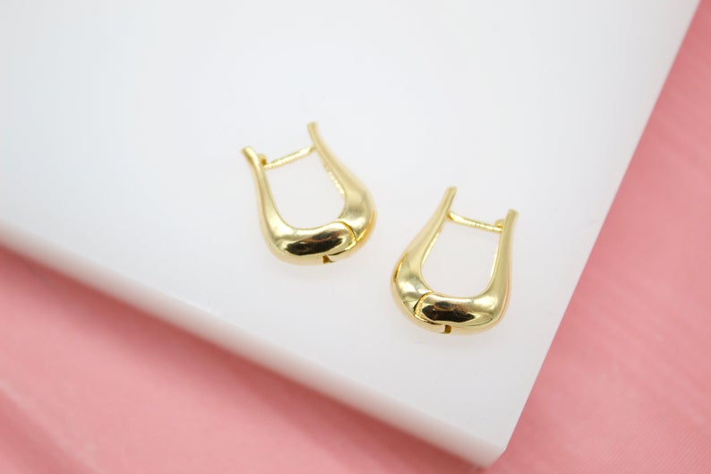A pair of polished gold U-shaped hoop earrings rests on a white surface against a pink background.