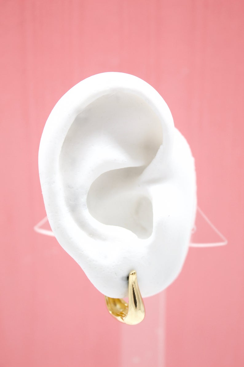A white model ear showcases a polished gold hoop earring against a soft pink background.