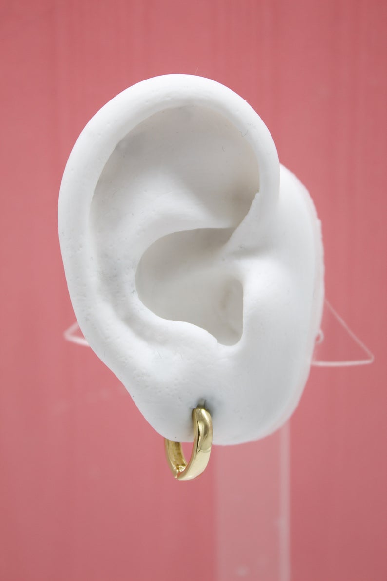 A white mannequin ear displays a small, polished gold huggie hoop earring against a solid pink backdrop.