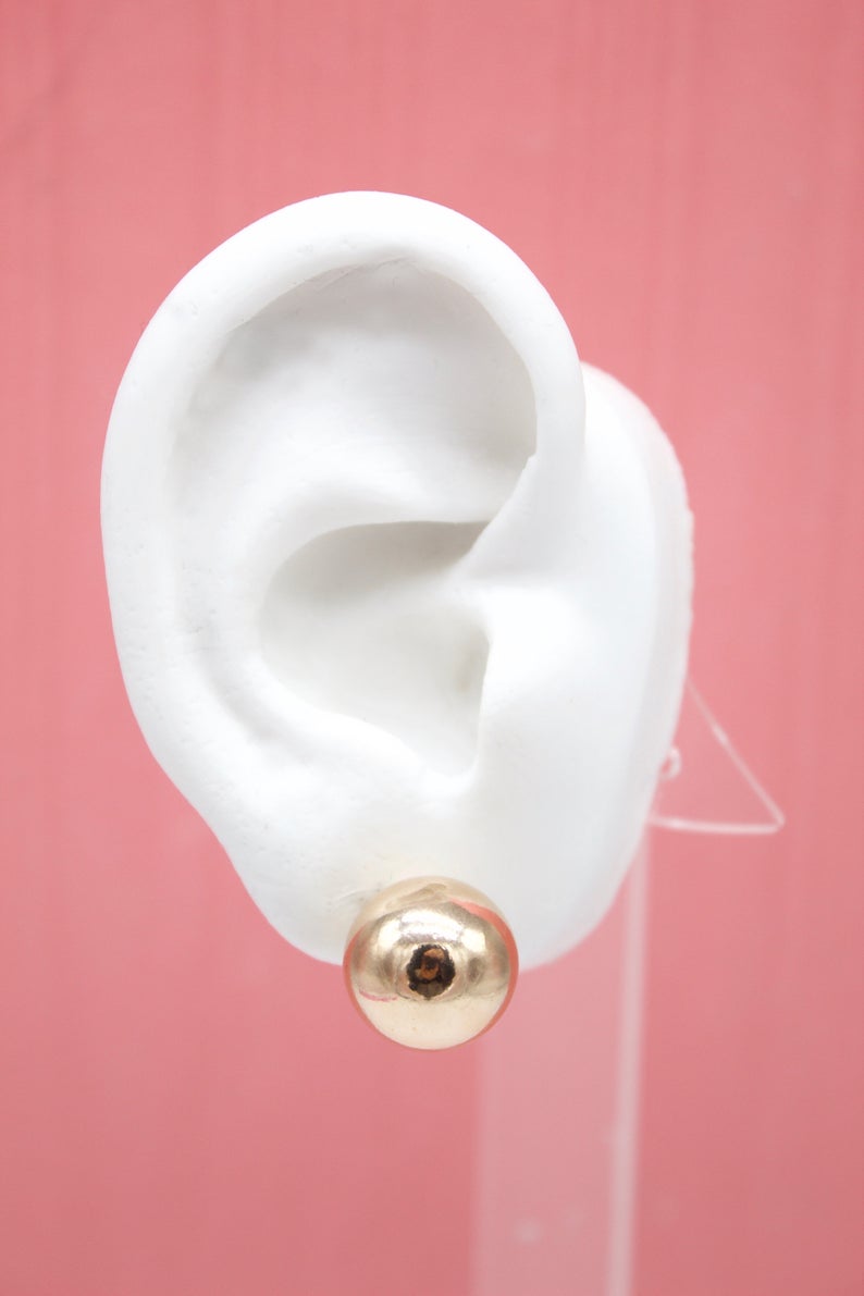 A white mannequin ear displays a polished gold ball stud earring against a pink background.