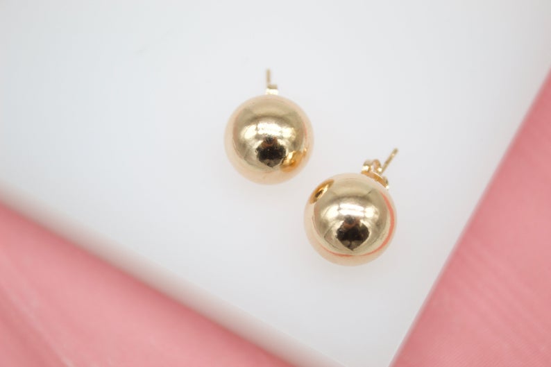 A pair of polished gold ball stud earrings rests on a white and pink display surface.