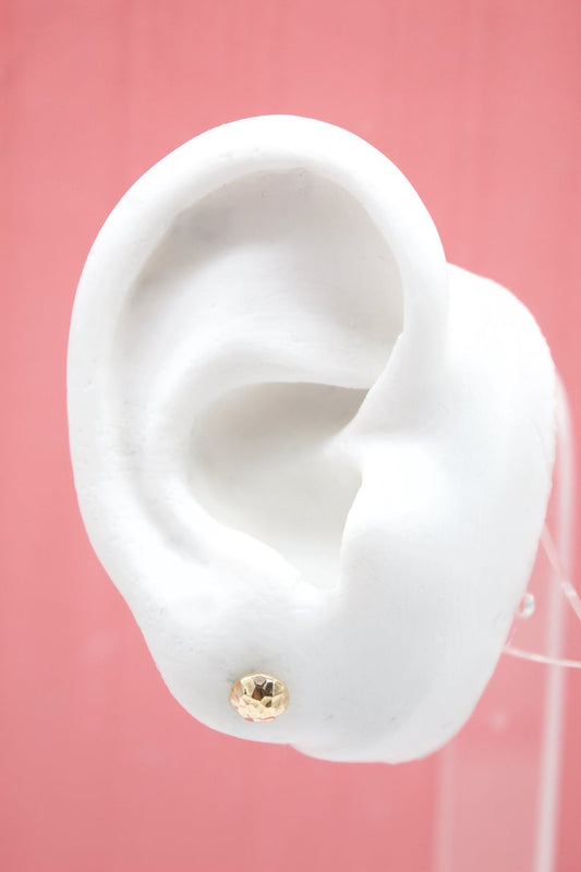 A white mannequin ear displays a small, faceted gold ball stud earring against a pink background.