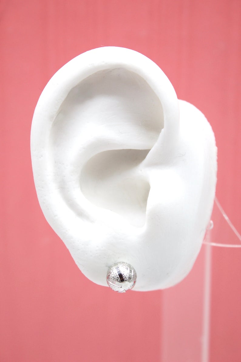 A white display ear model showcases a sparkling silver ball stud earring against a solid pink background.