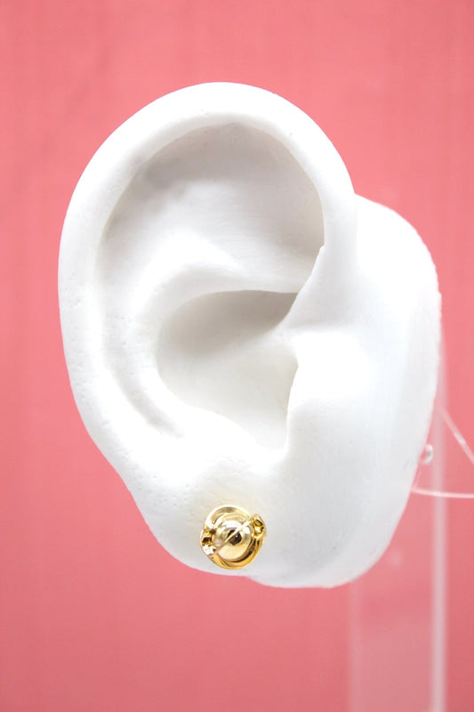 A white mannequin ear displays a shiny gold ball stud earring against a pink background.