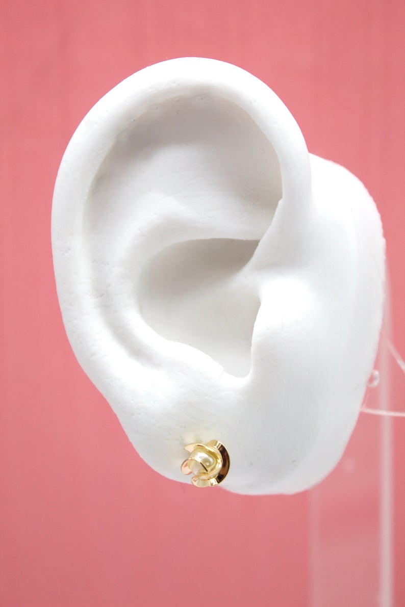 A white mannequin ear displays a shiny gold floral stud earring against a pink background.