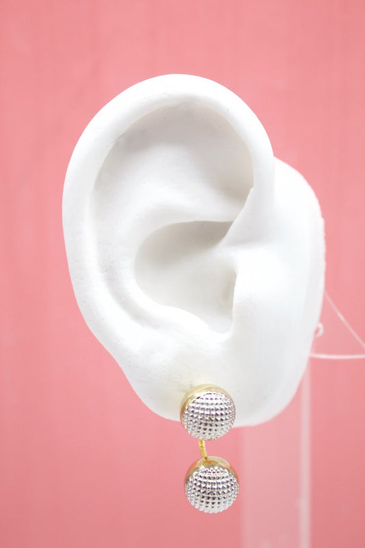 A white model ear showcases a double-sided gold stud earring with textured silver balls against a pink background.