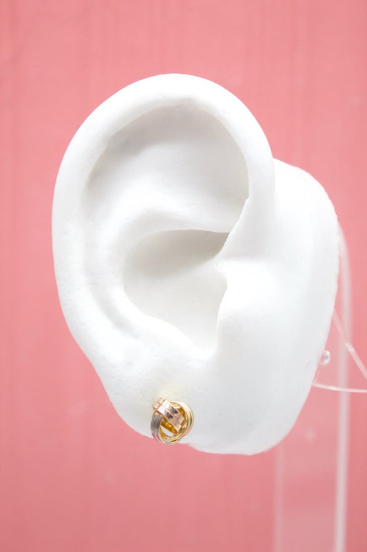 A white mannequin ear displays a small tricolor gold interlinked hoop stud earring against a pink background.