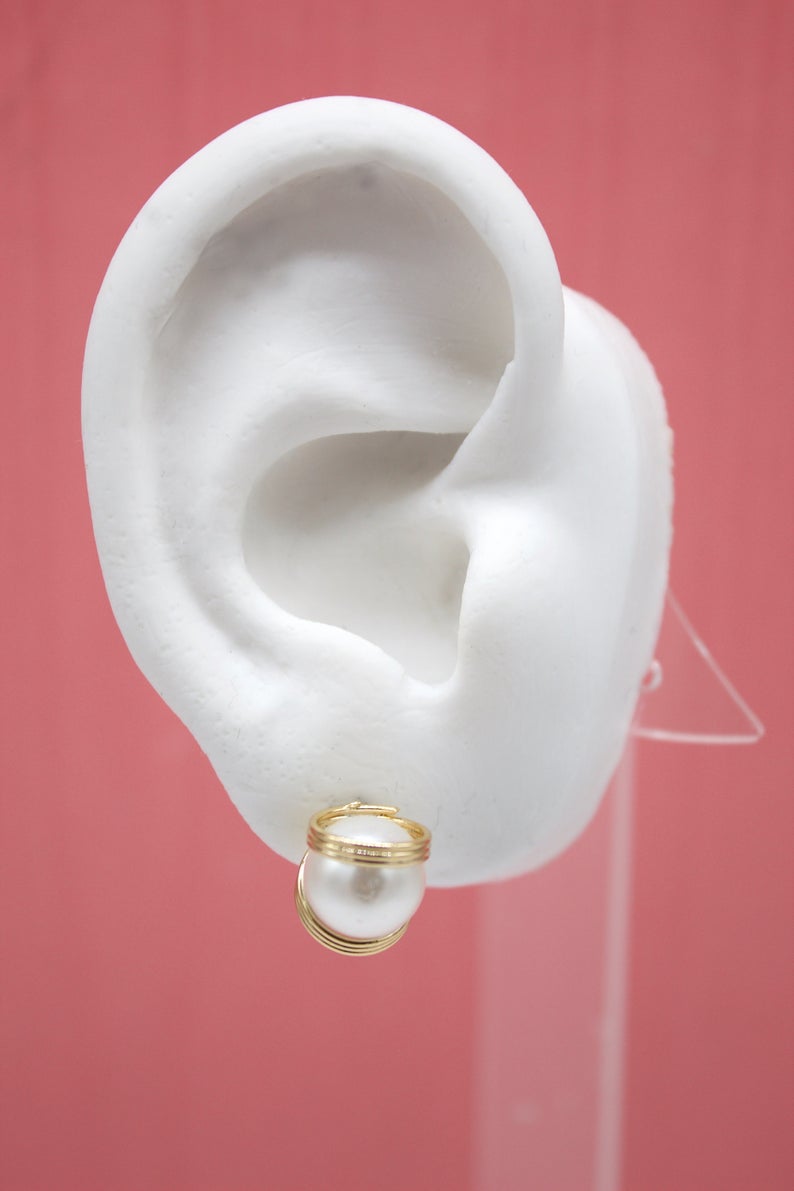 A white ear model displays a gold-wrapped pearl stud earring against a pink background.