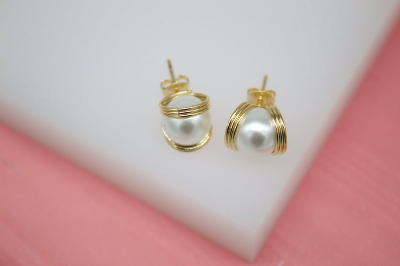 A pair of elegant gold-plated stud earrings featuring white pearls cradled in delicate wirework sits on a white surface.