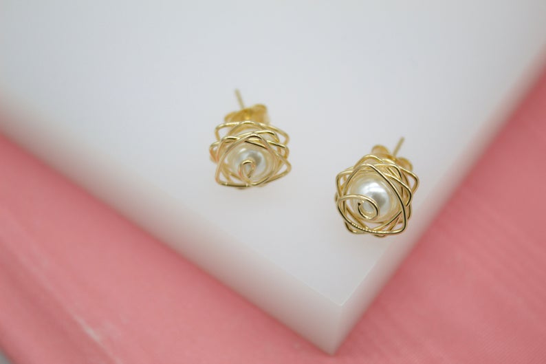 A pair of gold wire-wrapped stud earrings featuring lustrous white pearls rests on a white display block against a soft pink background.