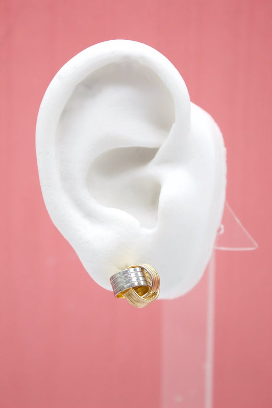 A white display mannequin ear showcases an intertwined gold and silver knot stud earring against a solid pink background.