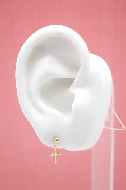 A white mannequin ear displays a delicate gold cross charm stud earring against a solid pink background.