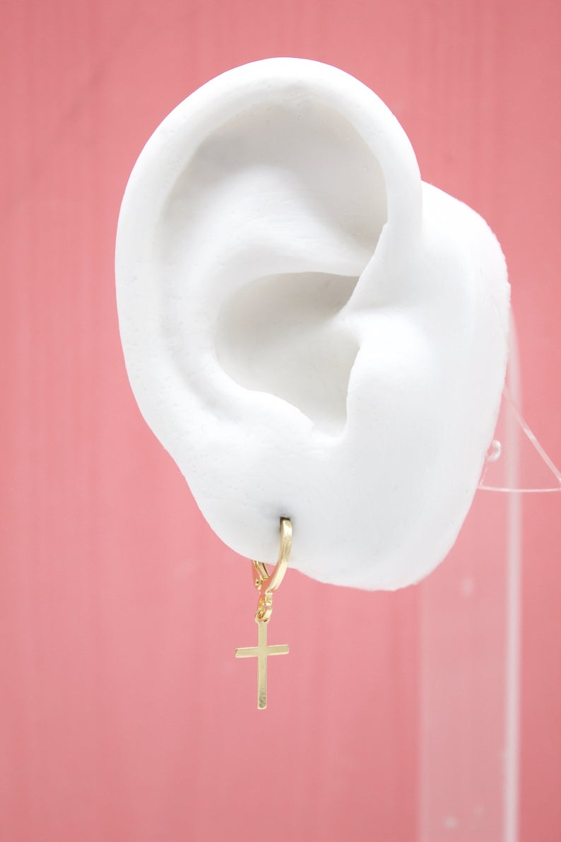A white mannequin ear displays a gold cross charm hoop earring against a pink background.