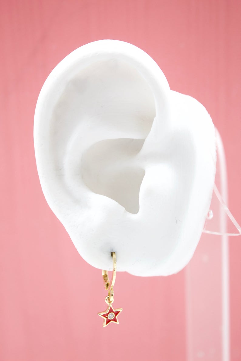 A white mannequin ear displays a gold huggie hoop earring featuring a red enamel star charm with a central stone.