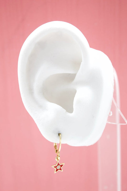 A white mannequin ear displays a gold huggie hoop earring featuring a red enamel star charm with a central stone.