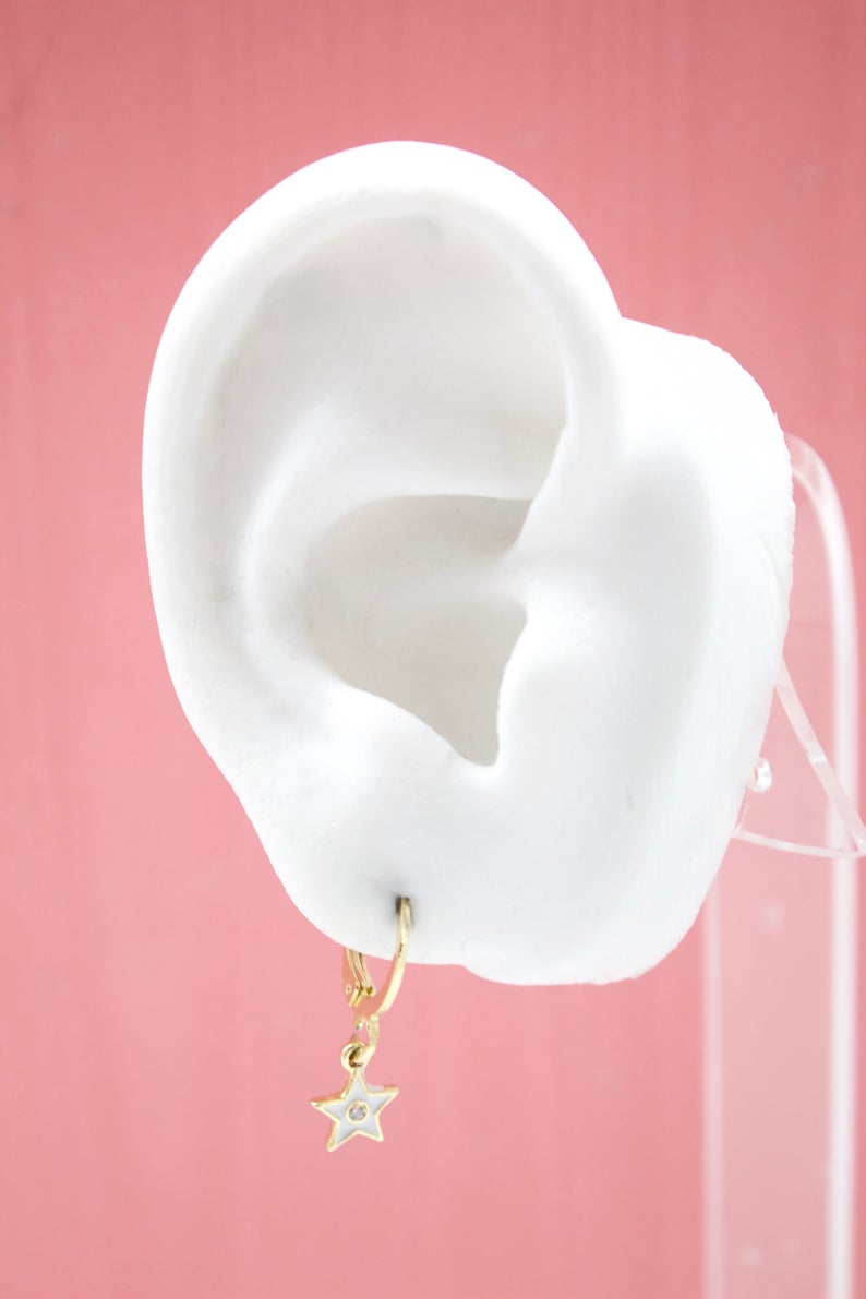 A white mannequin ear displays a gold star-shaped charm hoop earring against a pink background.