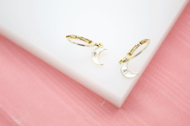 A pair of delicate gold crescent moon earrings featuring white enamel and sparkling crystal details rests on a white display surface against a pink background.