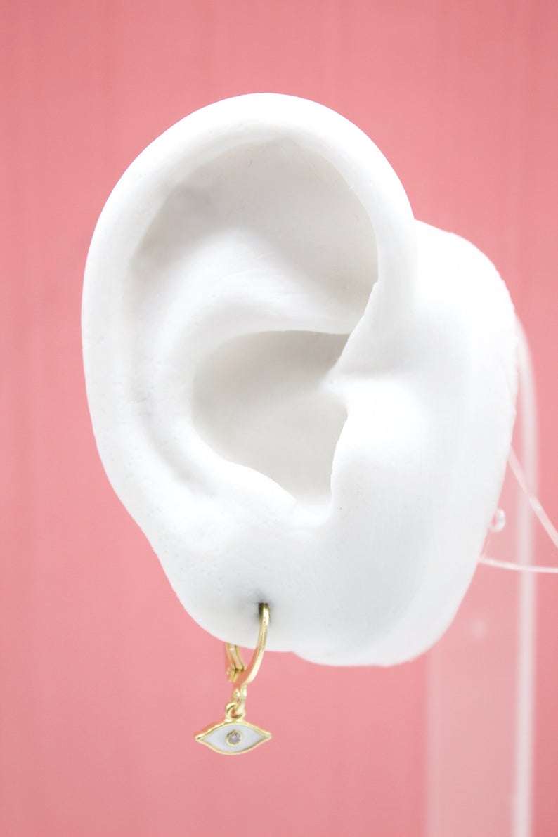 A white mannequin ear displays a gold evil eye charm huggie hoop earring against a pink background.