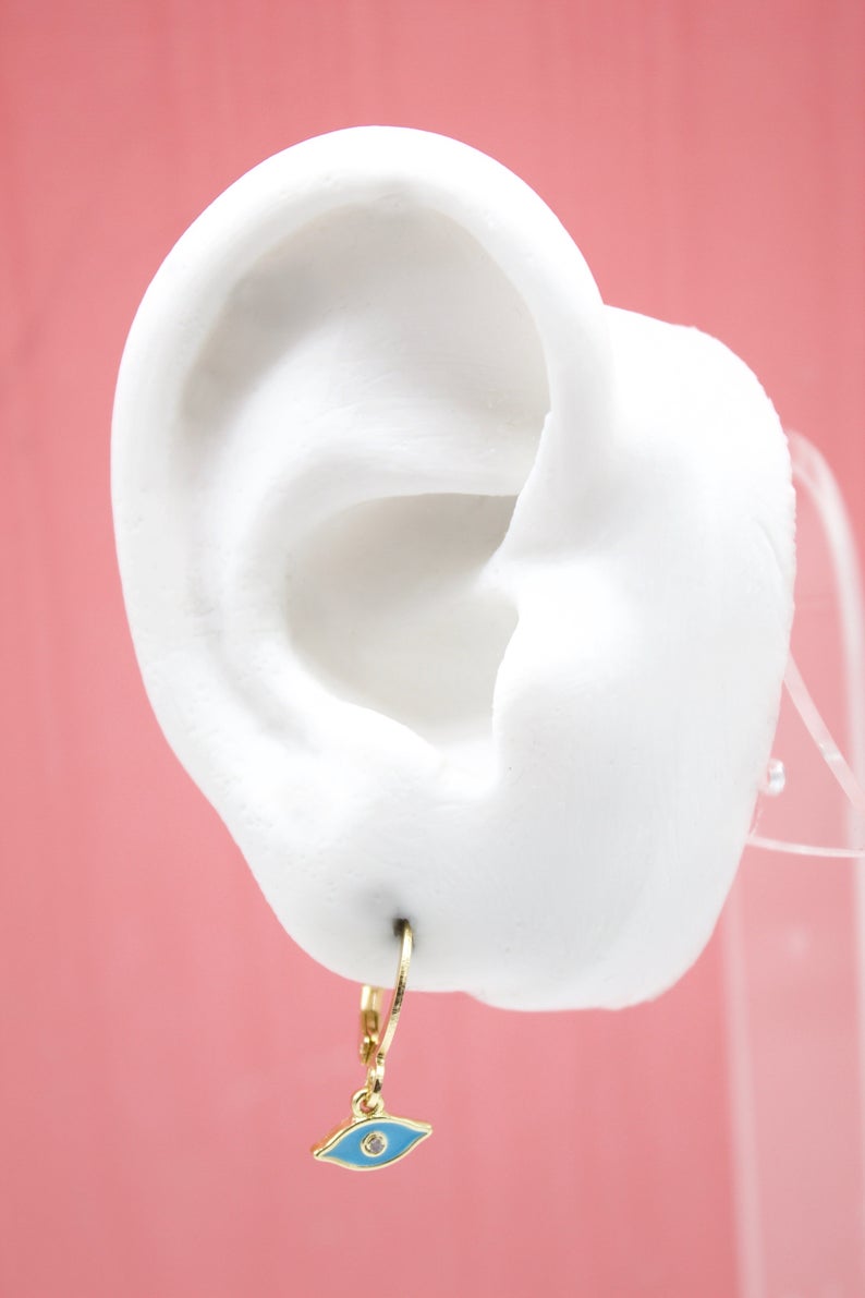 A white display ear model showcases a gold evil eye hoop earring with blue enamel against a pink background.