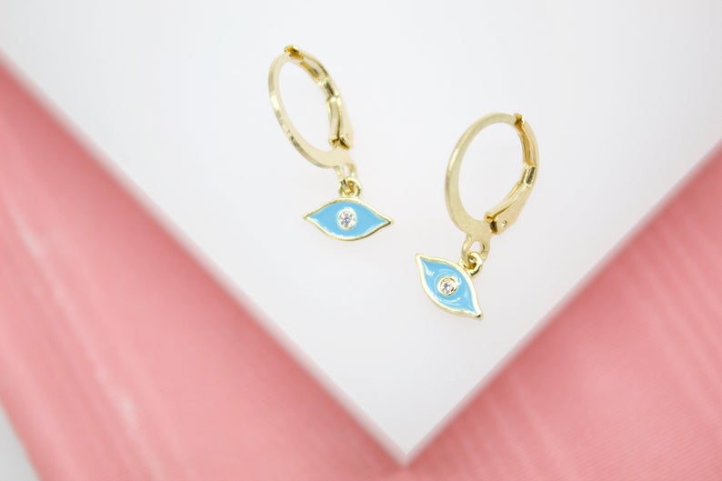 A pair of gold huggie hoop earrings featuring blue enamel evil eye charms with sparkling center stones rest on a white surface.