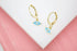 A pair of gold huggie hoop earrings featuring blue enamel evil eye charms with sparkling center stones rest on a white surface.