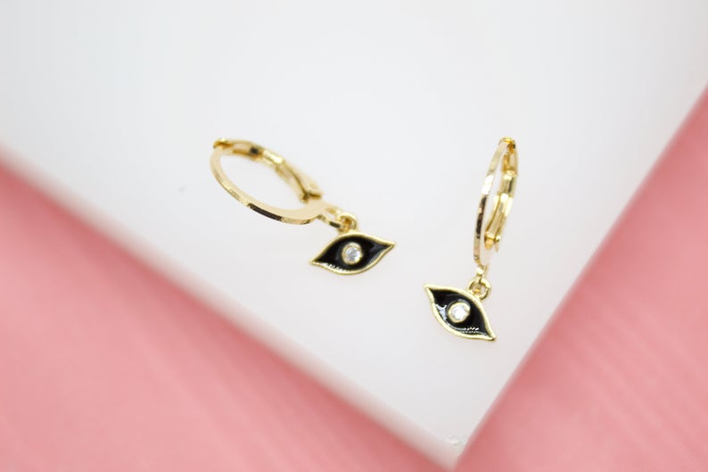 A pair of gold evil eye hoop earrings with black enamel and central cubic zirconia stones rest on a white surface.