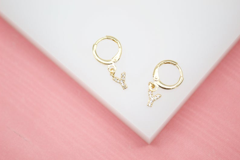 A pair of gold initial Y hoop earrings with sparkling crystal accents rests on a white and pink display surface.