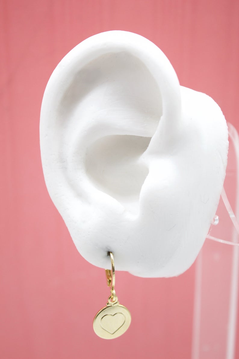 A white mannequin ear displays a gold huggie hoop earring with a round pendant featuring an engraved heart.