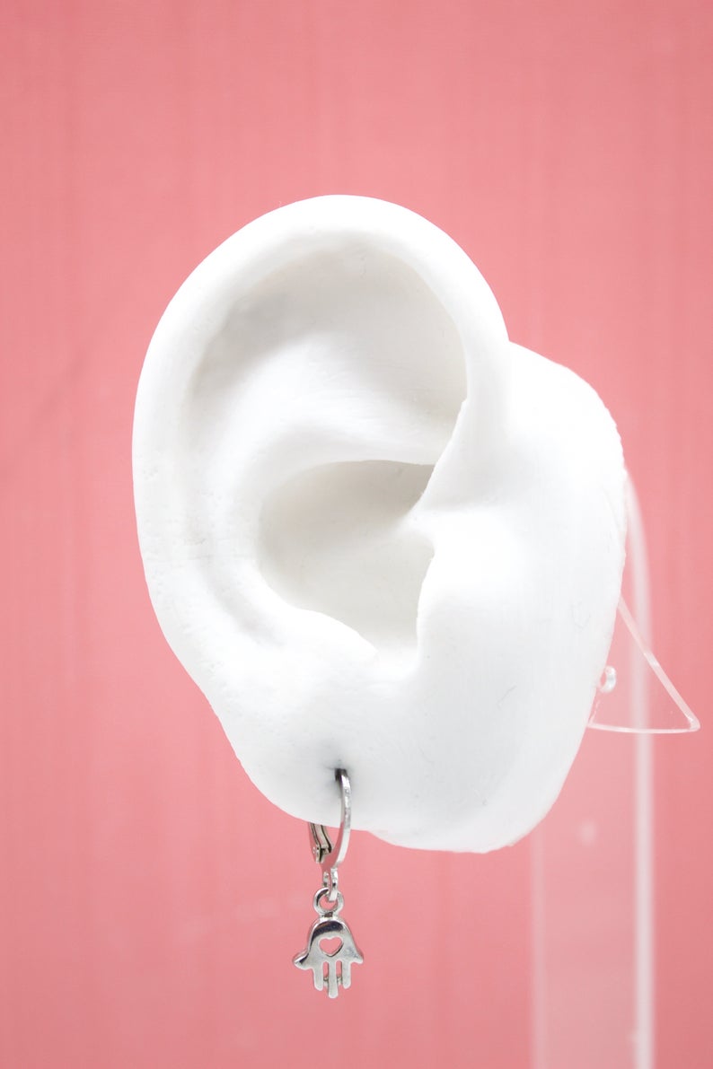 A white mannequin ear displays a silver hamsa hand charm hoop earring against a pink background.